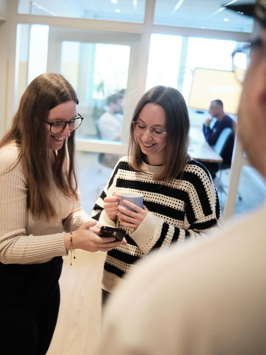 Austausch und Teamkultur – Kaffeepause bei der wemotion Digitalagentur Zwei Mitarbeiterinnen der wemotion Digitalagentur unterhalten sich lächelnd bei einer Kaffeepause und blicken auf ein Smartphone – Symbol für Austausch und Teamkultur.