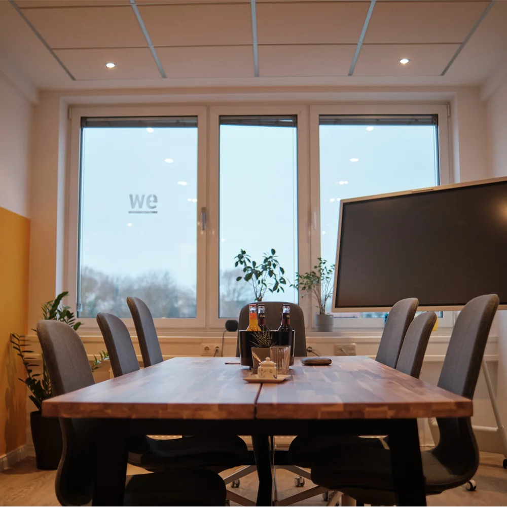 Besprechungsraum bei wemotion mit Holztisch, Fenstern und Bildschirm; mehrere Pflanzen im Raum.