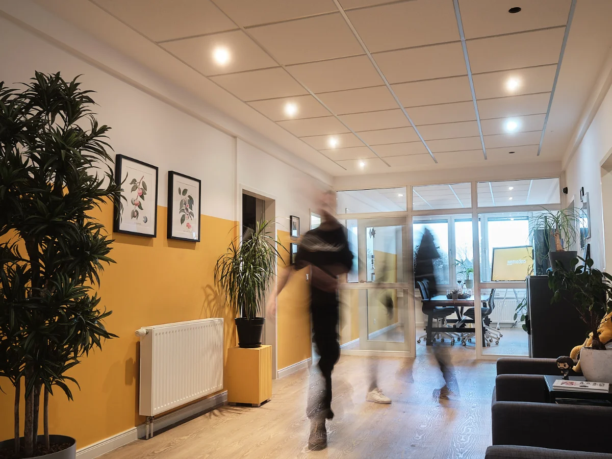 Büroflur der Digitalagentur wemotion mit gelber Akzentwand, gerahmten Bildern und mehreren großen Pflanzen; rechts Sofa, hinten verglaster Besprechungsraum; zwei vorbeigehende Personen in Bewegungsunschärfe.